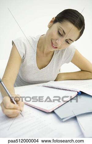 Picture - Woman sitting at desk, looking at document and writing in notebook. Fotosearch