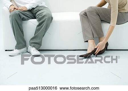 Stock Photograph - Man and woman sitting on sofa, woman putting on shoe, cropped view. Fotosearch