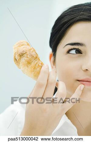 Stock Image - Woman holding croissant, looking away, cropped. Fotosearch