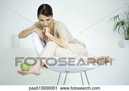 Stock Image - Woman sitting in chair, balancing apple on foot. Fotosearch