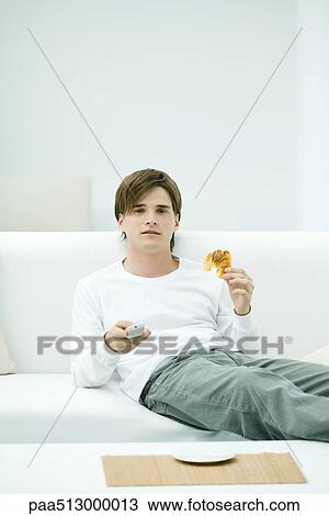 Young man sitting on sofa, holding remote control and croissant View Large Photo Image Stock Image - Young man sitting on sofa, holding remote control and croissant. Fotosearch