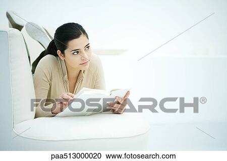 Young woman lying on stomach on sofa, holding book, looking away View Large Photo Image Stock Image - Young woman lying on stomach on sofa, holding book, looking away. Fotosearch