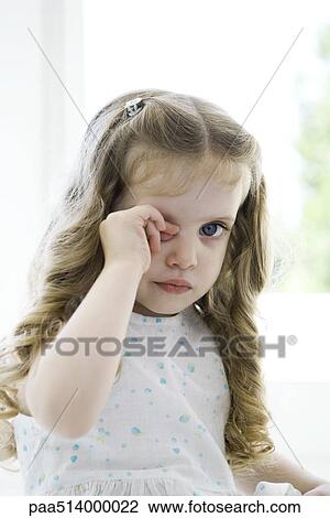 Stock Image - Little girl rubbing her eye, looking at camera. Fotosearch