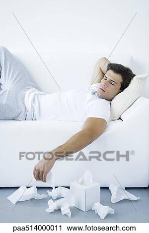 Stock Image - Man lying on sofa, discarded tissues on the floor beside him. Fotosearch