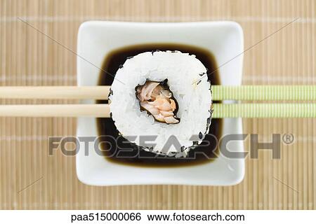 Stock Photograph - Maki sushi resting on chopsticks above soy sauce, overhead view. Fotosearch