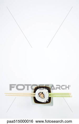 Stock Photograph - Single piece of maki sushi on chopsticks over dish of soy sauce, overhead view. Fotosearch