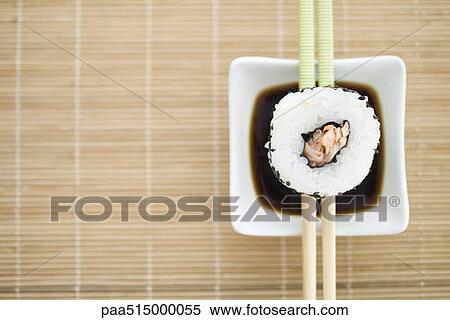 Stock Photography - Single piece of maki sushi resting on chopsticks over sauce, overhead view. Fotosearch