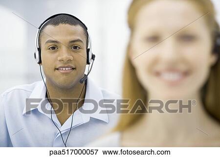 Stock Photo - Professional man wearing headset smiling at camera, female colleague in foreground. Fotosearch
