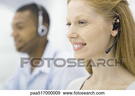 Stock Photo - Woman wearing headset, male colleague in background, both working, smiling. Fotosearch