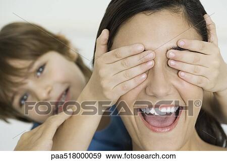 Stock Photo - Boy covering mother's eyes with his hands, woman laughing. Fotosearch