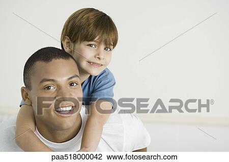 Stock Image - Boy leaning over father's shoulders, both smiling, portrait. Fotosearch