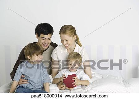 Parents and two children sitting together on bed, one boy holding gift View Large Photo Image Picture - Parents and two children sitting together on bed, one boy holding gift. Fotosearch