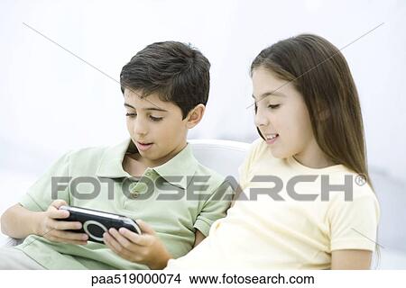 Brother and sister sitting together, boy playing handheld video game View Large Photo Image Picture - Brother and sister sitting together, boy playing handheld video game. Fotosearch