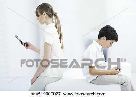 Young woman holding cell phone with back turned on boy playing handheld video game View Large Photo Image Stock Photo - Young woman holding cell phone with back turned on boy playing handheld video game. Fotosearch