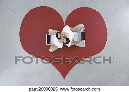 Stock Image - Couple sitting back to back on large heart, holding laptop computers, smiling up at camera. Fotosearch
