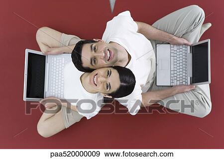 Couple sitting back to back on the ground, holding laptop computers, smiling up at camera View Large Photo Image Stock Photo - Couple sitting back to back on the ground, holding laptop computers, smiling up at camera. Fotosearch