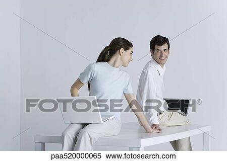 Stock Photography - Couple sitting back to back with laptops on their laps, holding hands, smiling at each other. Fotosearch
