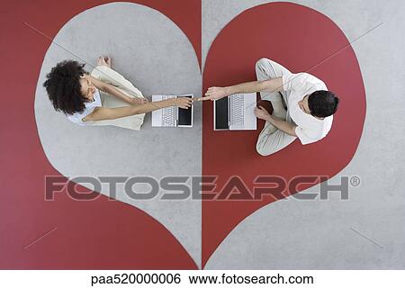 Stock Photograph - Couple sitting face to face with laptop computers on heart shape, touching fingers, overhead view. Fotosearch