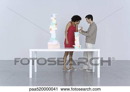 Stock Image - Couple standing beside table stacked with boxes, man giving woman a gift. Fotosearch