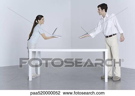 Man and woman at opposite ends of table, holding laptop computers toward one another View Large Photo Image Stock Image - Man and woman at opposite ends of table, holding laptop computers toward one another. Fotosearch