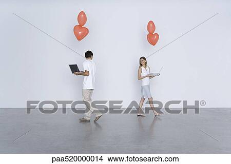 Picture - Man and woman carrying laptop computers and heart balloons, smiling over their shoulders at each other. Fotosearch