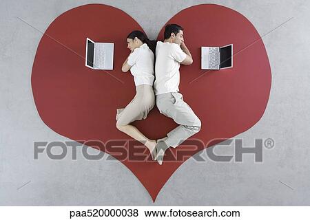 Stock Photo - Man and woman lying back to back on large heart, both looking at laptop computers. Fotosearch