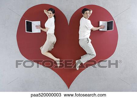 Stock Image - Man and woman lying on large heart graphic, both using laptop computers. Fotosearch