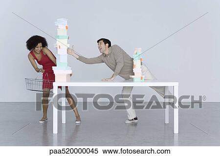 Stock Photography - Man and woman reaching for tall stack of boxes, woman holding shopping basket. Fotosearch