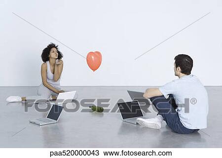 Stock Image - Man and woman seated with laptops, woman blowing man a kiss, bottle and heart balloon between them. Fotosearch