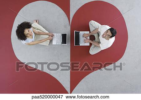 Stock Photo - Man and woman sitting on large heart, using laptop computers, smiling up at camera. Fotosearch