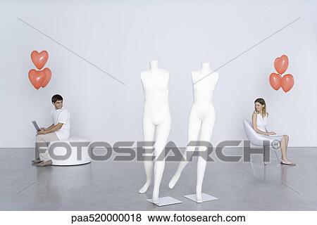 Stock Photo - Man and woman sitting with laptops and heart balloons, looking over shoulders at each other. Fotosearch