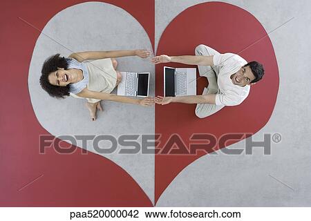 Stock Image - Man and woman sitting with laptops on large heart, arms out, touching hands, overhead view. Fotosearch
