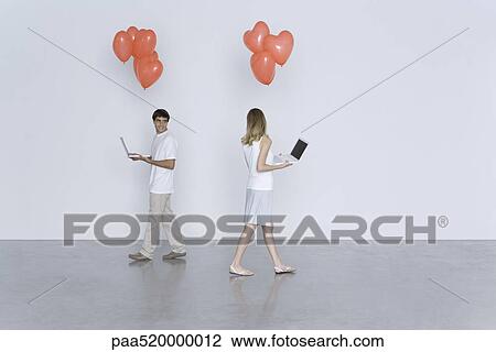 Stock Image - Man and woman walking past each other, both carrying laptop computers and heart balloons. Fotosearch
