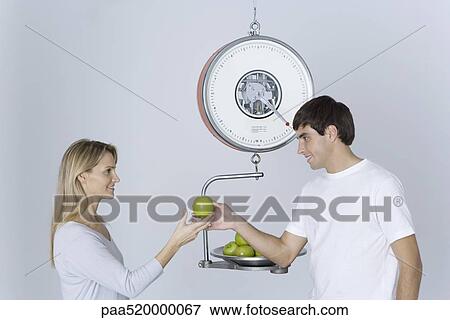 Stock Photo - Man weighing apples on scale, giving woman one apple. Fotosearch
