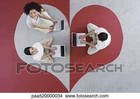 Two women and one man sitting on large heart, using laptop computers, women smiling up at camera View Large Photo Image Picture - Two women and one man sitting on large heart, using laptop computers, women smiling up at camera. Fotosearch