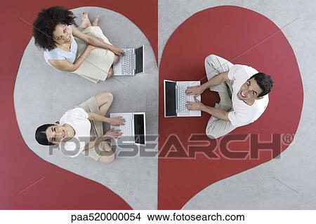 Picture - Two women and one man sitting on large heart, using laptop computers, smiling up at camera. Fotosearch