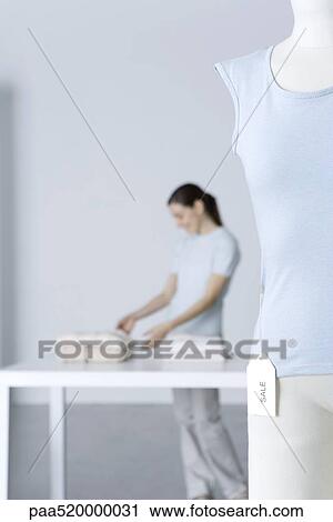 Stock Image - Woman looking at clothing folded on table, focus on tee-shirt with "sale" tag in foreground. Fotosearch