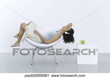 Stock Image - Woman reclining in chair, listening to headphones connected to apple, side view. Fotosearch