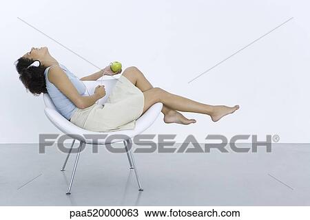 Woman reclining in chair, listening to headphones connected to apple, side view View Large Photo Image Stock Image - Woman reclining in chair, listening to headphones connected to apple, side view. Fotosearch