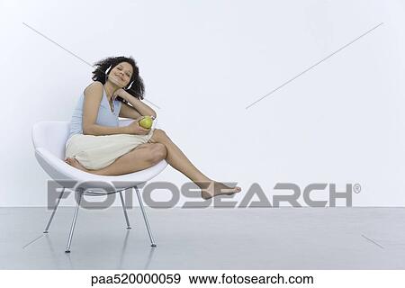 Woman sitting in chair, listening to headphones connected to apple, smiling View Large Photo Image Stock Photo - Woman sitting in chair, listening to headphones connected to apple, smiling. Fotosearch