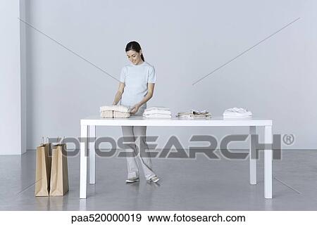 Stock Photo - Woman standing at table, looking at folded sweater. Fotosearch