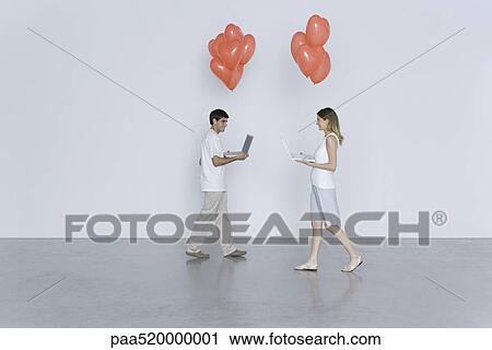 Stock Image - Young man and woman walking toward each other, both carrying laptop computers and heart balloons. Fotosearch