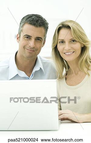 Picture - Couple sitting together with laptop, smiling at camera. Fotosearch