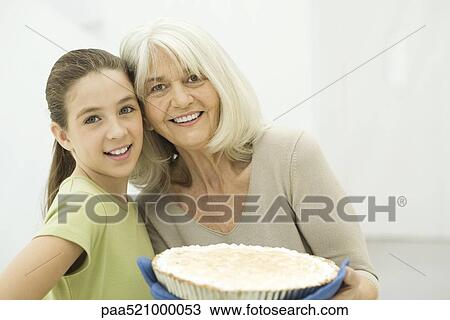 Stock Image - Grandmother and granddaughter smiling at camera, woman holding pie. Fotosearch