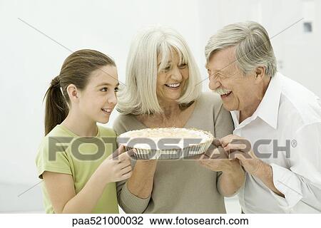 Stock Image - Grandparents and granddaughter smiling at each other, woman holding pie. Fotosearch
