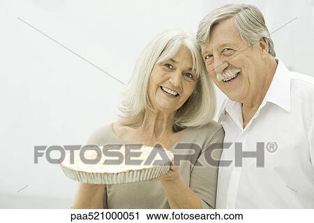 Stock Image - Senior couple smiling, woman holding up pie. Fotosearch