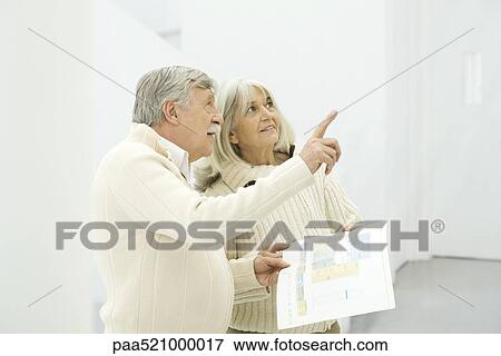 Stock Photo - Senior couple standing together, man holding floor plans and pointing up. Fotosearch