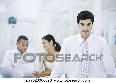 Businessman in office, smiling at camera, employees working in background View Large Photo Image Stock Image - Businessman in office, smiling at camera, employees working in background. Fotosearch