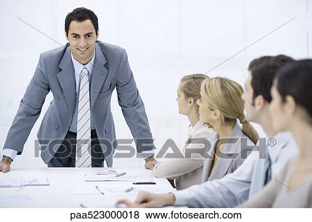 Businessman leaning over table addressing colleagues, smiling at camera View Large Photo Image Stock Photo - Businessman leaning over table addressing colleagues, smiling at camera. Fotosearch