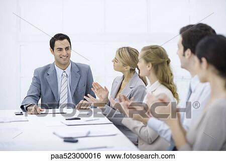 Stock Photograph - Businessman seated at head of table, smiling, colleagues clapping. Fotosearch
